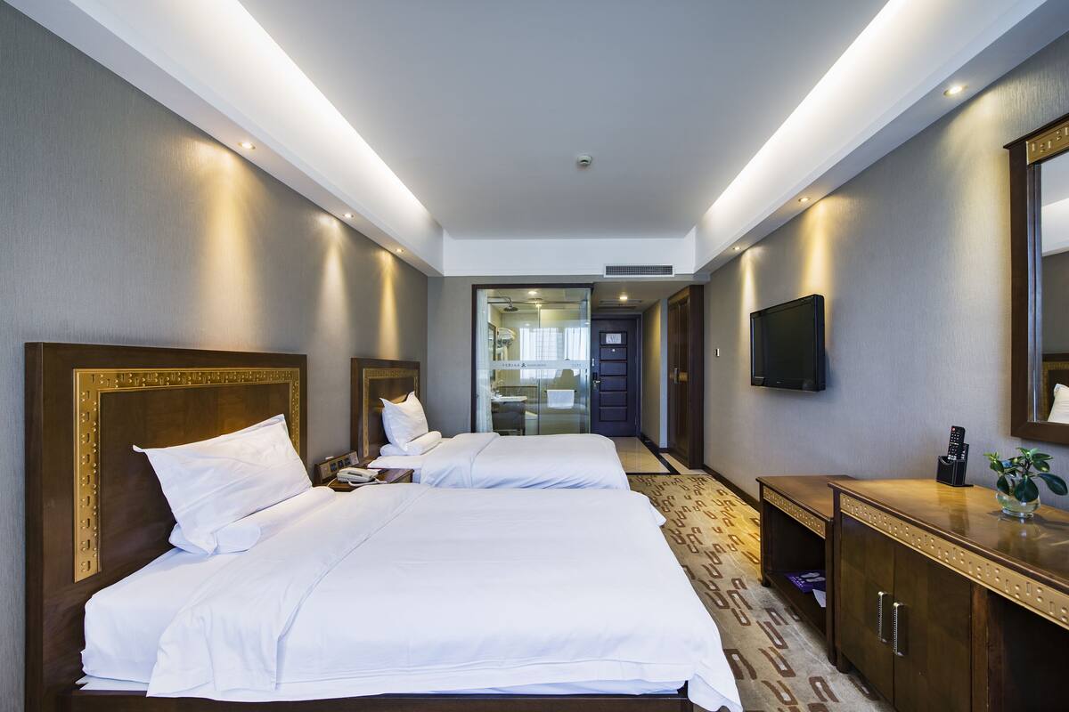 Habitación de hotel con dos camas, televisión, escritorio con una planta y baño visible a través de una puerta de vidrio.