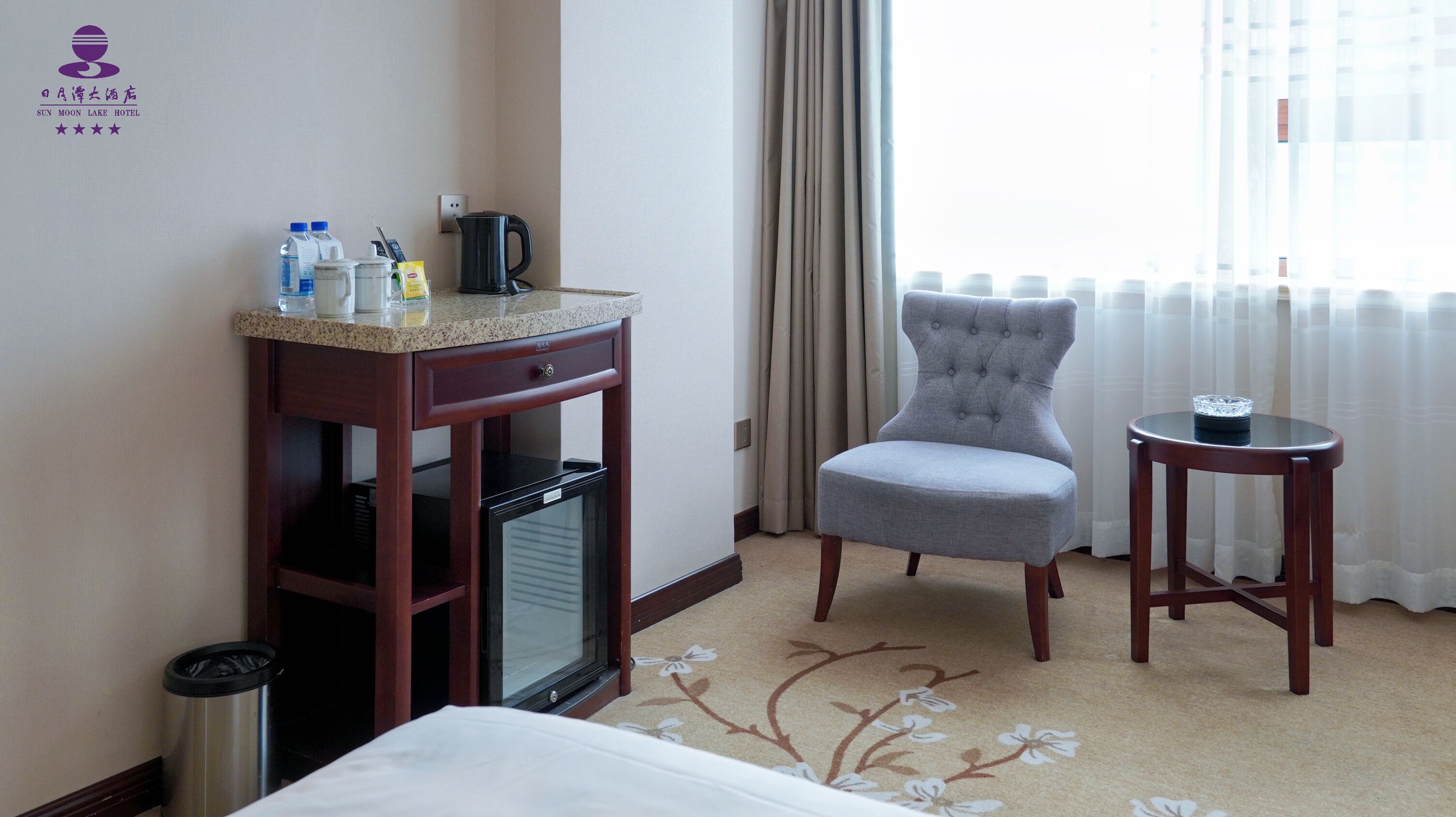 A hotel room with a bed, a small desk with a kettle, a chair, a small table, and a trash can.