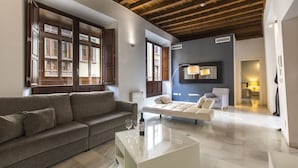 A modern living room with a sofa, a coffee table with wine glasses, and a large window with wooden shutters. - tuAnfitrion Elvira Suites (Granada)