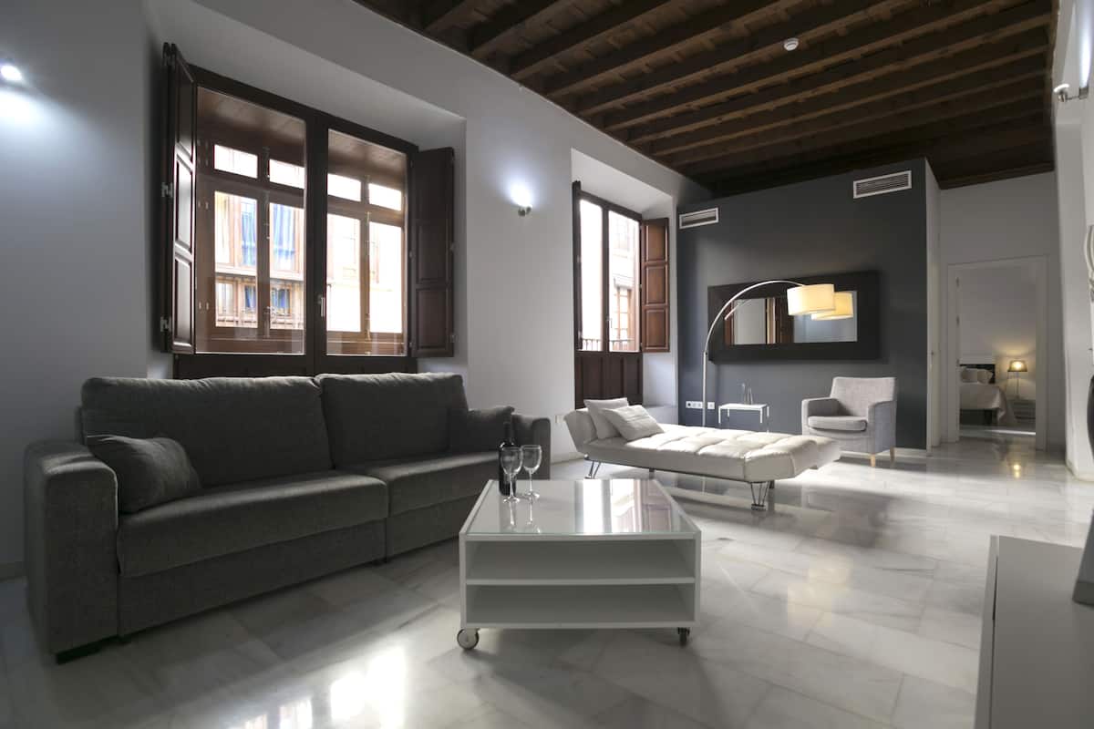 A modern living room with a grey sofa, a white coffee table, and a white armchair. There are large windows with wooden shutters, a flat-screen TV mounted on the wall, and a ceiling with exposed wooden beams.