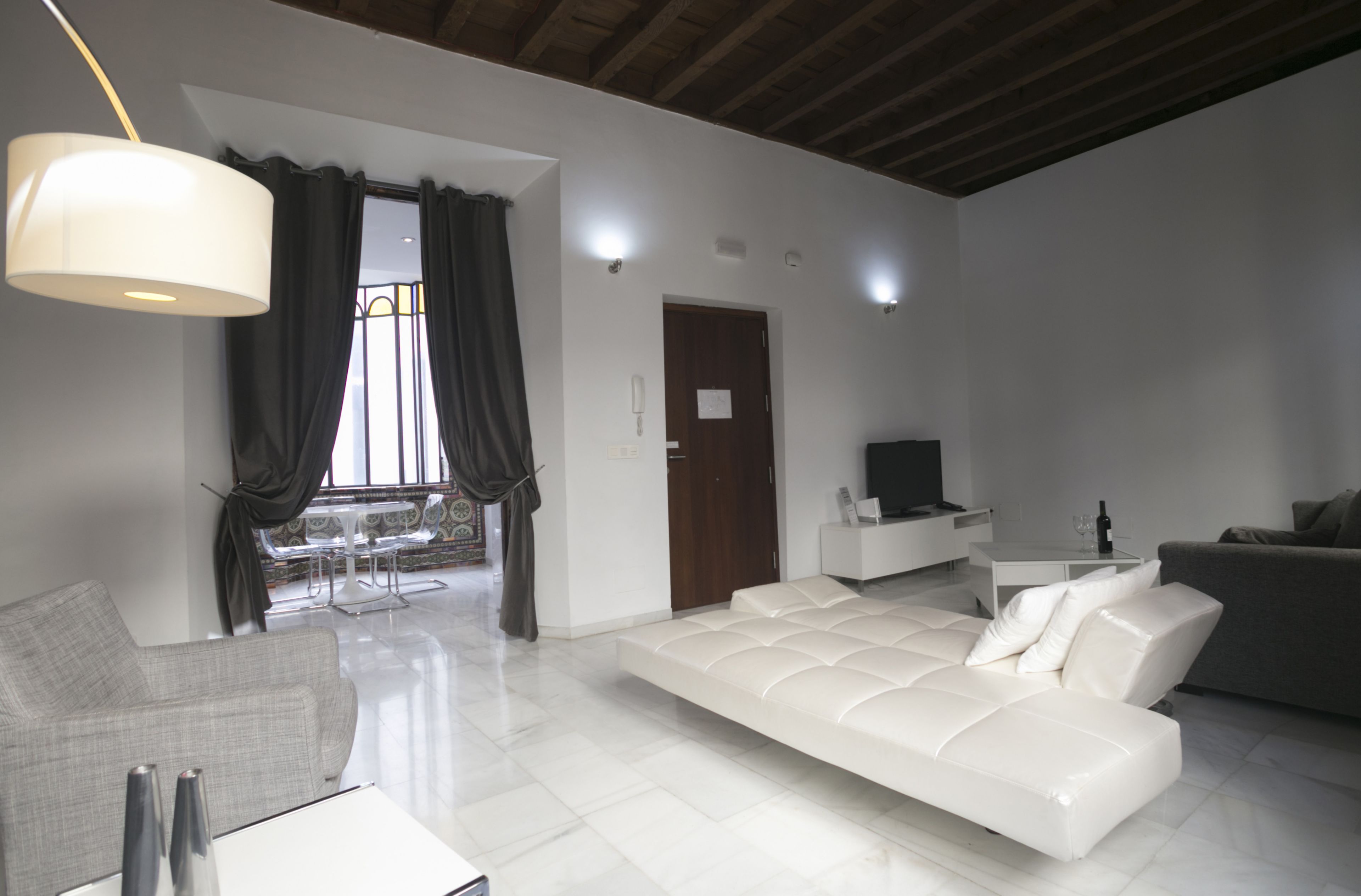Un salon moderne avec un canapé blanc, un fauteuil gris, une table basse blanche et un meuble pour la télévision. On y trouve une grande fenêtre avec des rideaux, un luminaire au plafond et une porte.