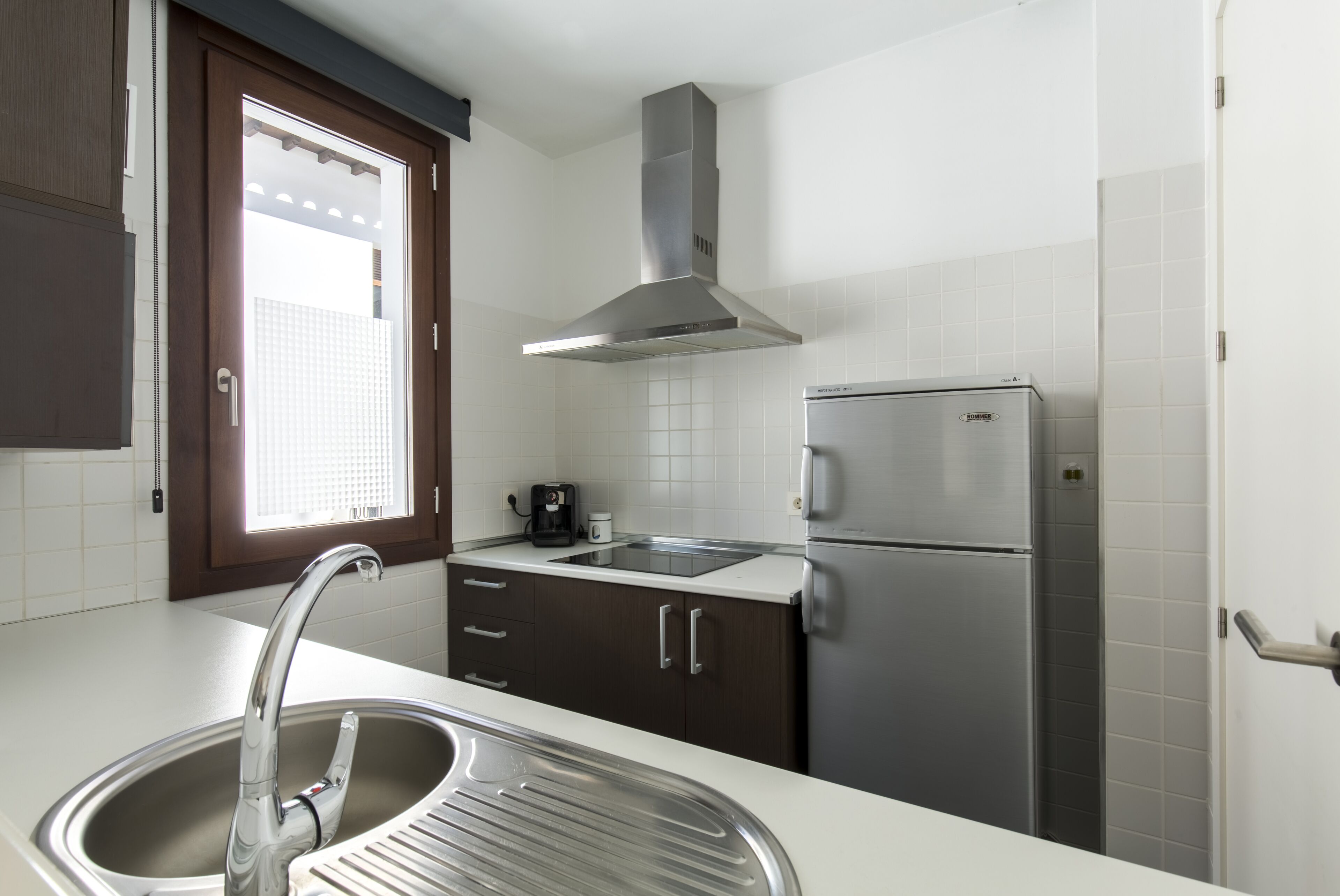 Eine Küche mit einem Edelstahl-Kühlschrank, einem Einbauofen, einer Kaffeemaschine und einem Fenster mit Jalousien.