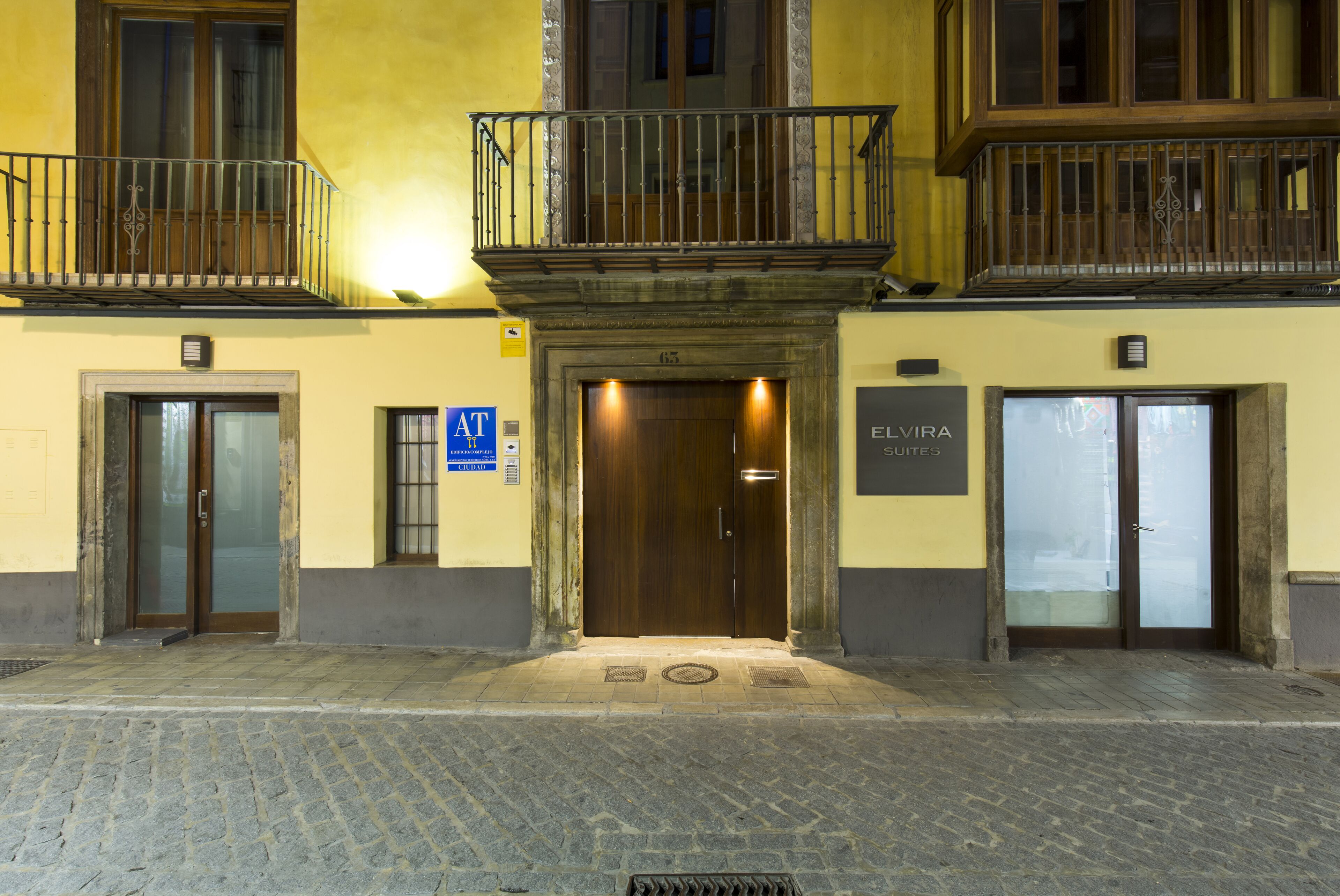 A building with a wooden entrance door, a sign for 'ELVIRA Suites', and a blue 'AT' sign on the left.