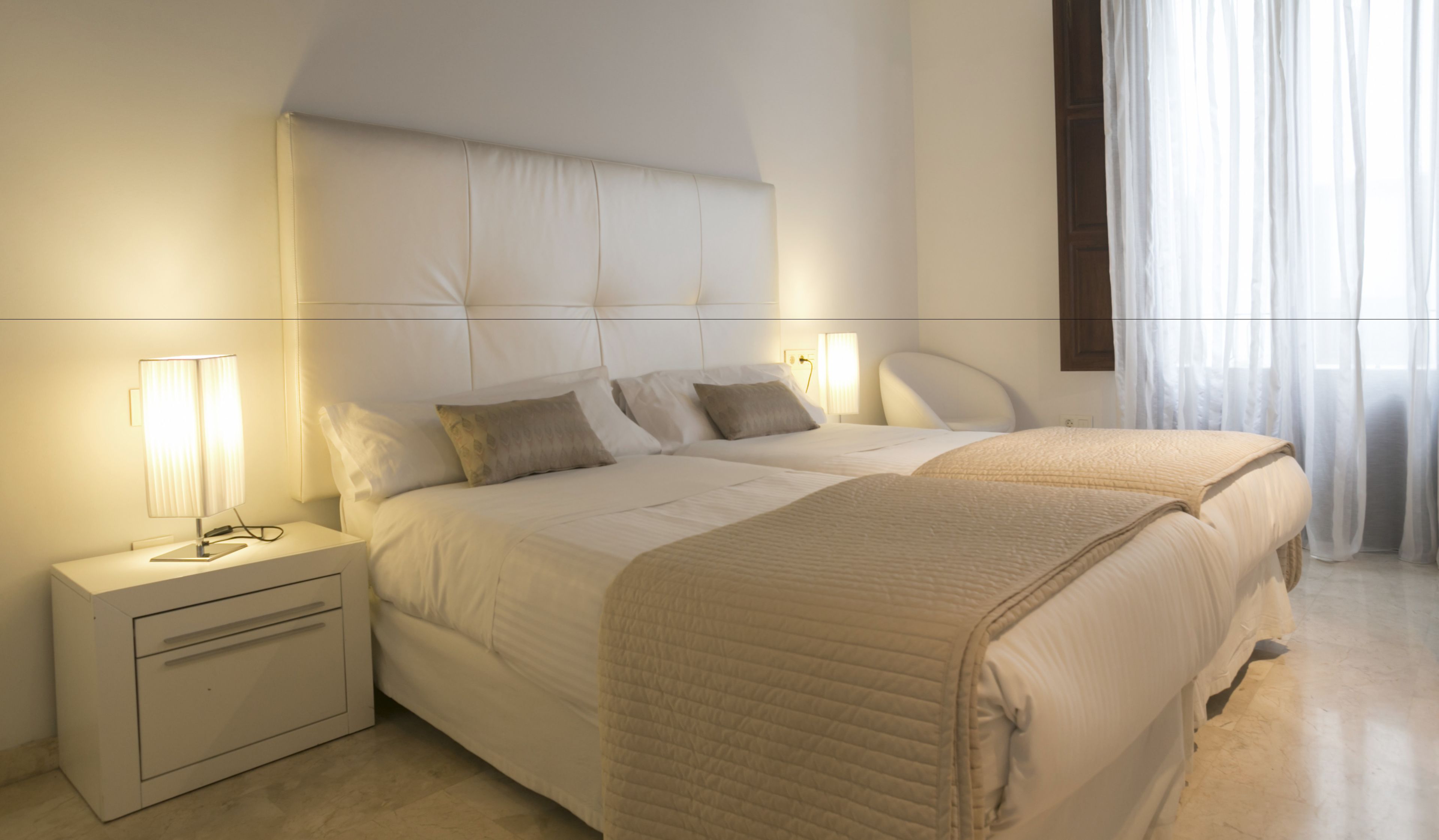 A hotel room with a large bed, two bedside tables, a nightstand, and a window with sheer curtains.