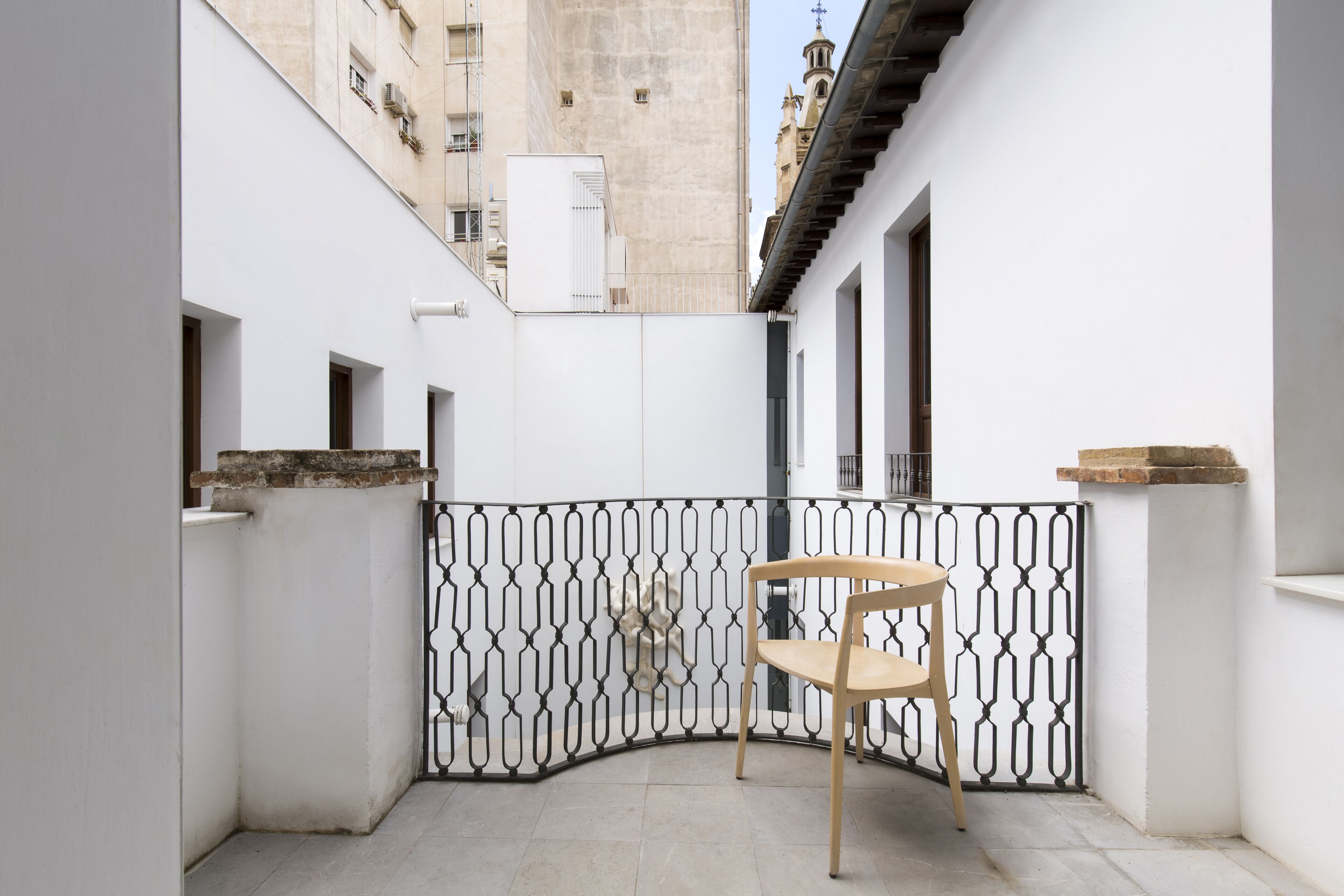 Un patio estrecho con una silla de madera, paredes blancas y una verja decorativa de metal.