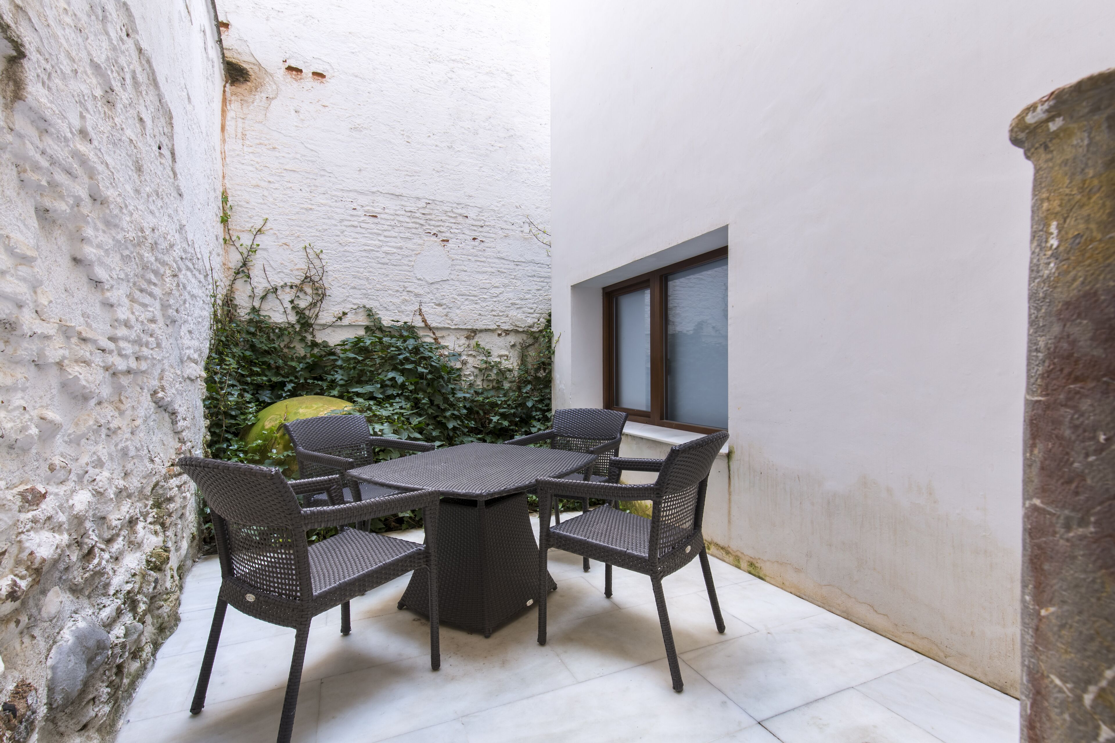 En hyggelig udendørs terrasse med et bord og stole, omgivet af en hvid mur og grøn vegetation.