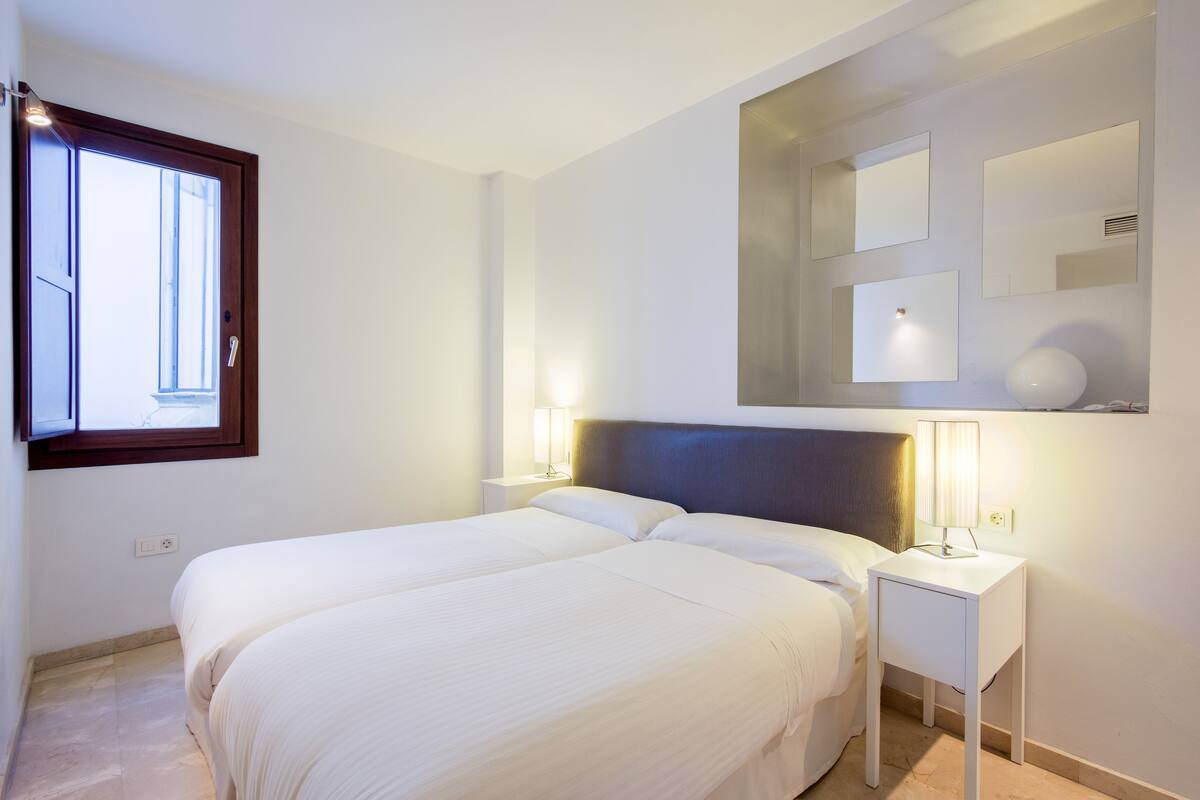 A bedroom with two beds, a nightstand with a lamp, a window, and a wall-mounted mirror.