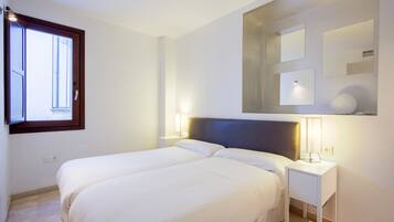 A bedroom with two beds, a nightstand with a lamp, a window, and a wall-mounted mirror.