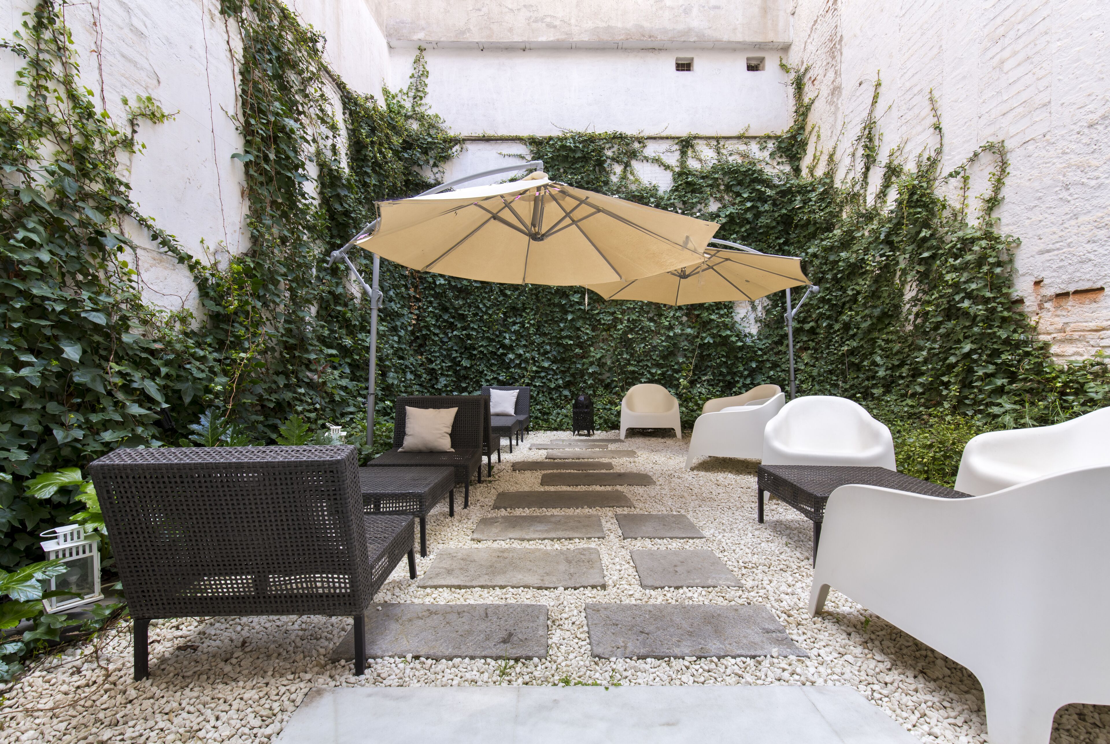 An outdoor seating area with wicker furniture and a beige umbrella, surrounded by a gravel path and lush greenery.