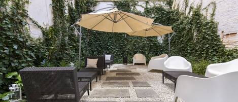 An outdoor seating area with wicker furniture and a beige umbrella, surrounded by a gravel path and lush greenery.