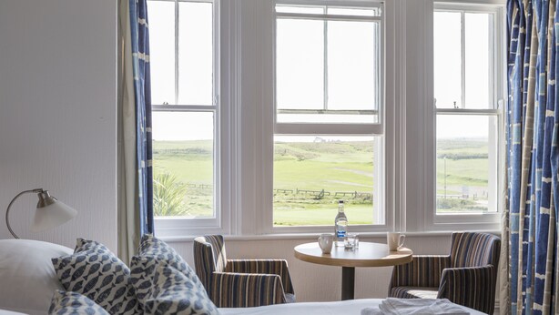 Habitación de hotel con una cama, dos sillas a rayas, una mesita con una botella y una taza, y vista a un paisaje verde a través de grandes ventanales.