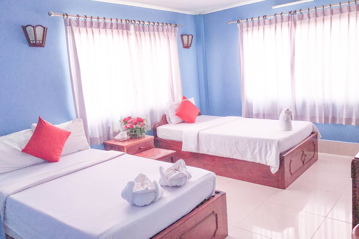 Une chambre d’hôtel avec deux lits, chacun agrémenté de draps blancs et d’oreillers rouges, une table de chevet avec un vase de fleurs et une commode en bois.