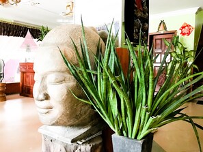 Une pièce comprenant un grand buste sculpté en pierre, une plante en pot aux longues feuilles vertes et un meuble en bois sur lequel repose un objet décoratif rouge.