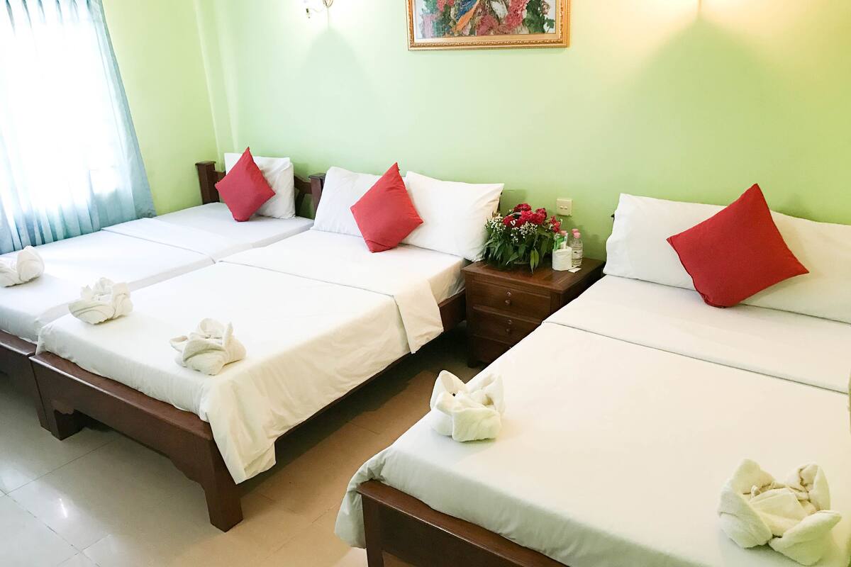 Une chambre d’hôtel avec trois lits, chacun étant équipé de draps blancs et d’oreillers rouges. On y trouve une table de chevet avec un vase de fleurs et des articles de toilette.