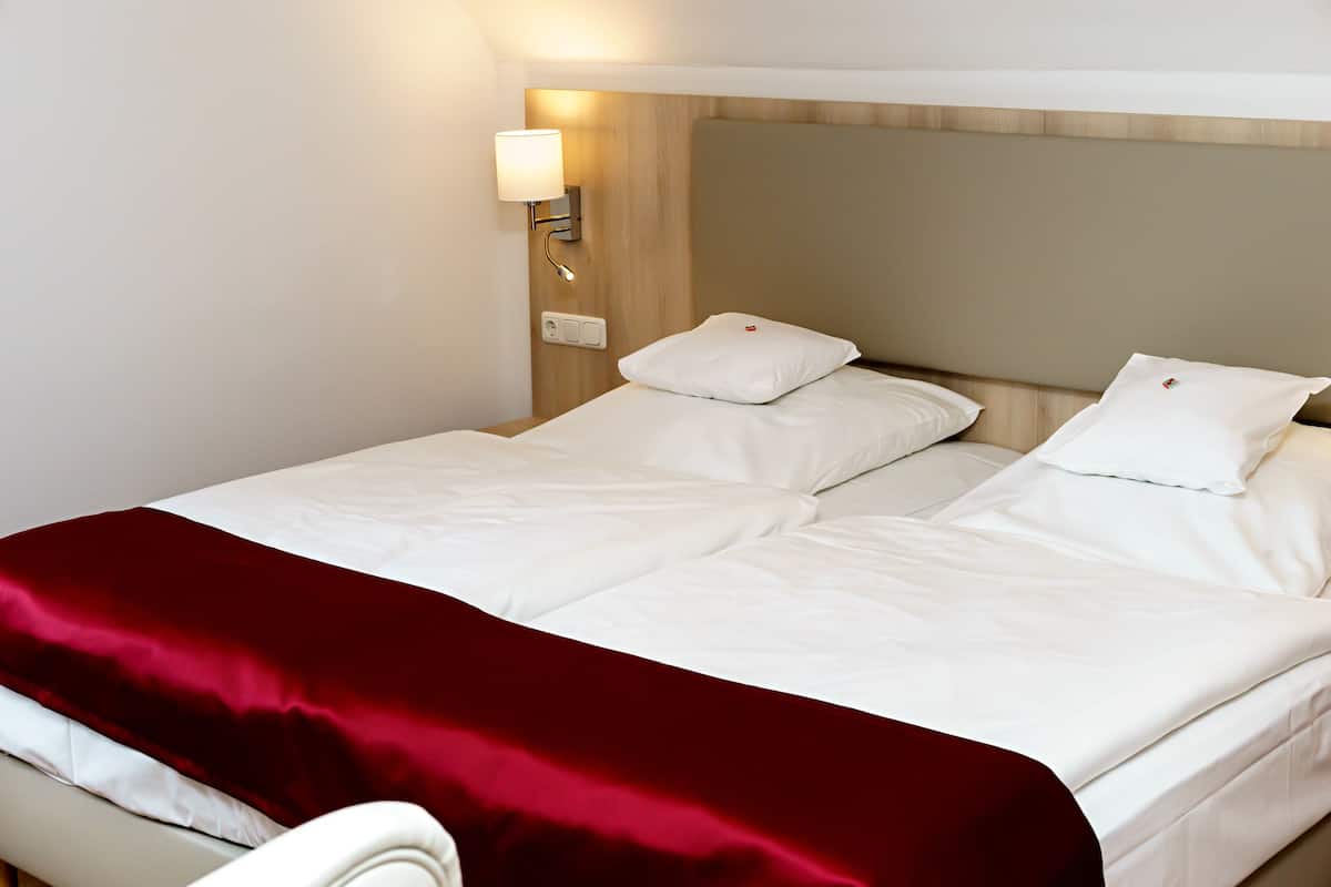 Quarto de hotel com cama, roupa de cama branca, manta vermelha e luminária fixada na parede.