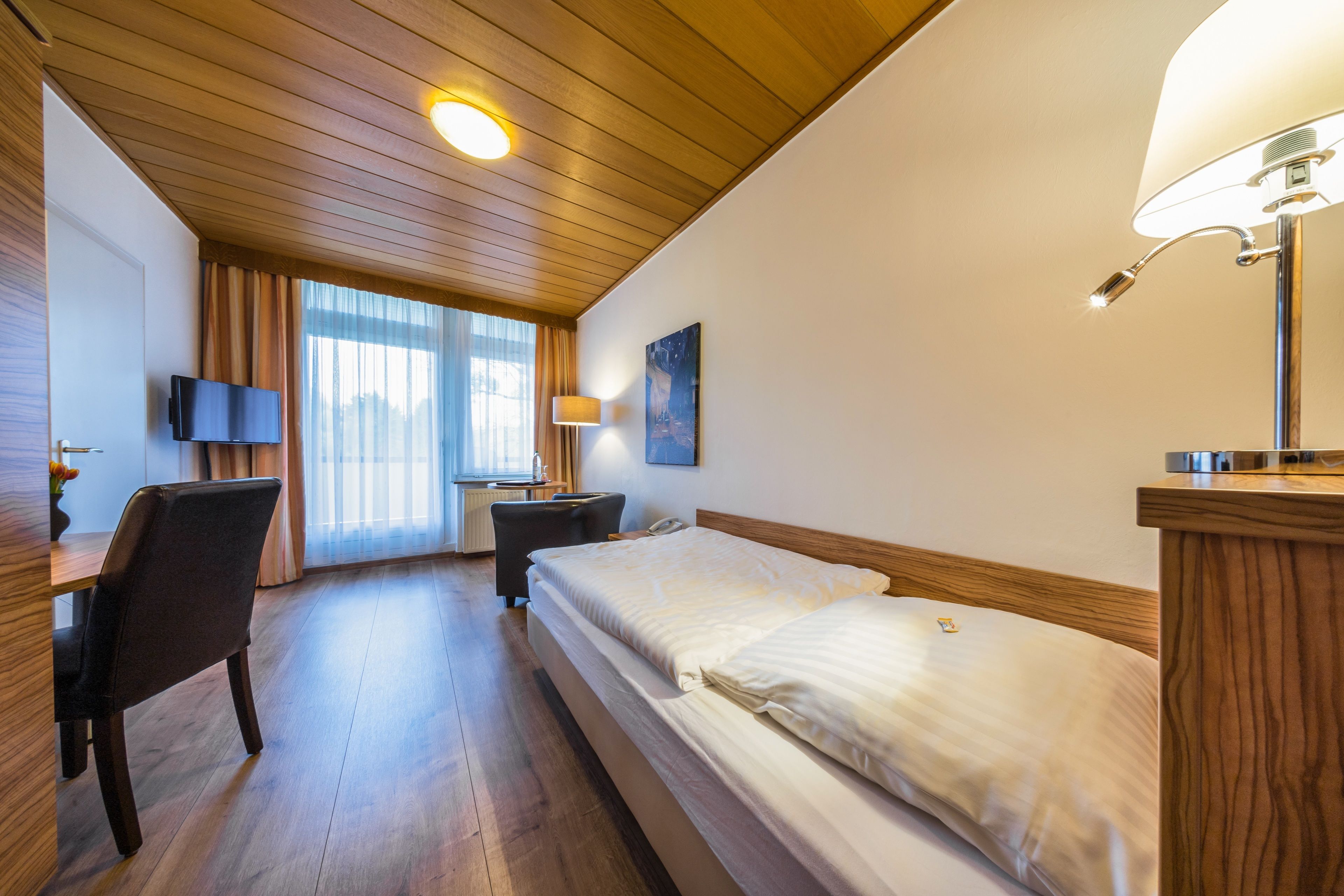 Habitación de hotel con techo de madera, una cama con ropa de cama blanca, un escritorio con silla, un televisor y una lámpara.