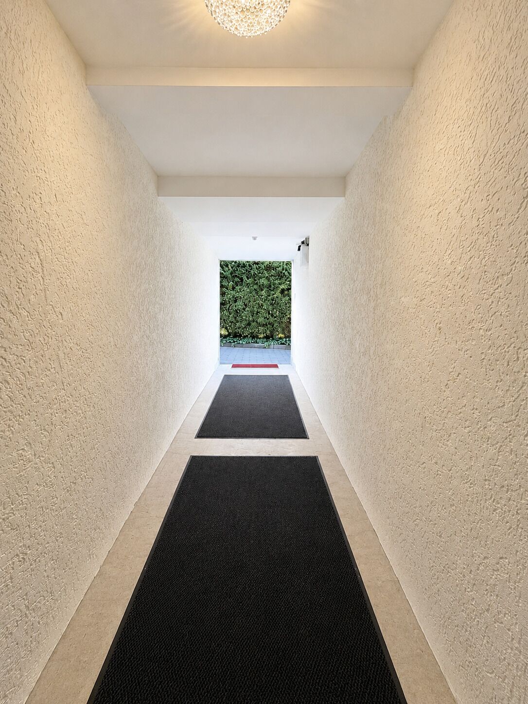 A hallway with textured walls, black carpets, and a view of a green hedge outside.