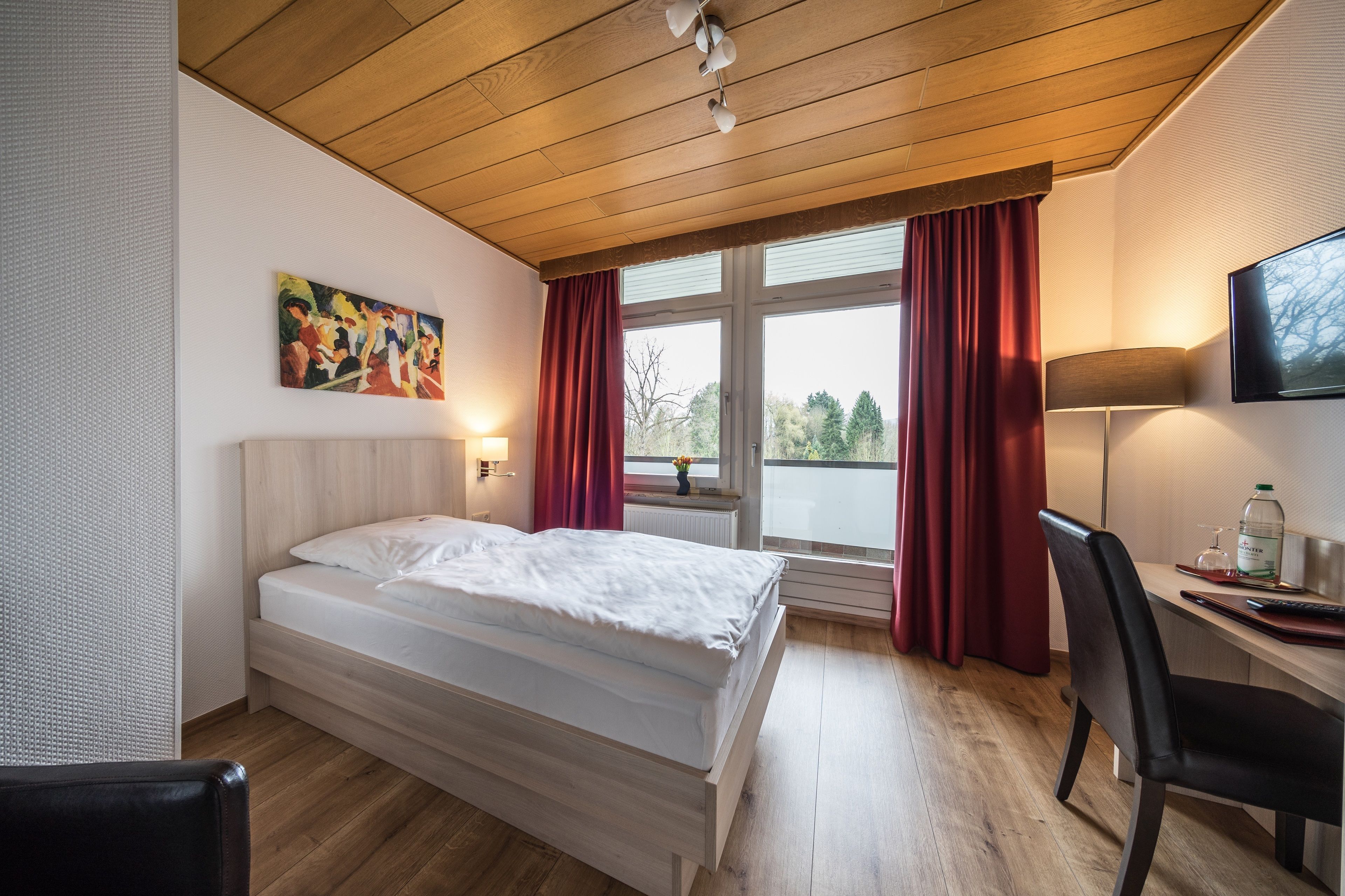 Hotelkamer met een bed, bureau, stoel, raam met gordijnen, een schilderij aan de muur en een aan de muur bevestigde televisie.