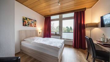 Hotelkamer met een bed, bureau, stoel, raam met gordijnen, een schilderij aan de muur en een aan de muur bevestigde televisie.
