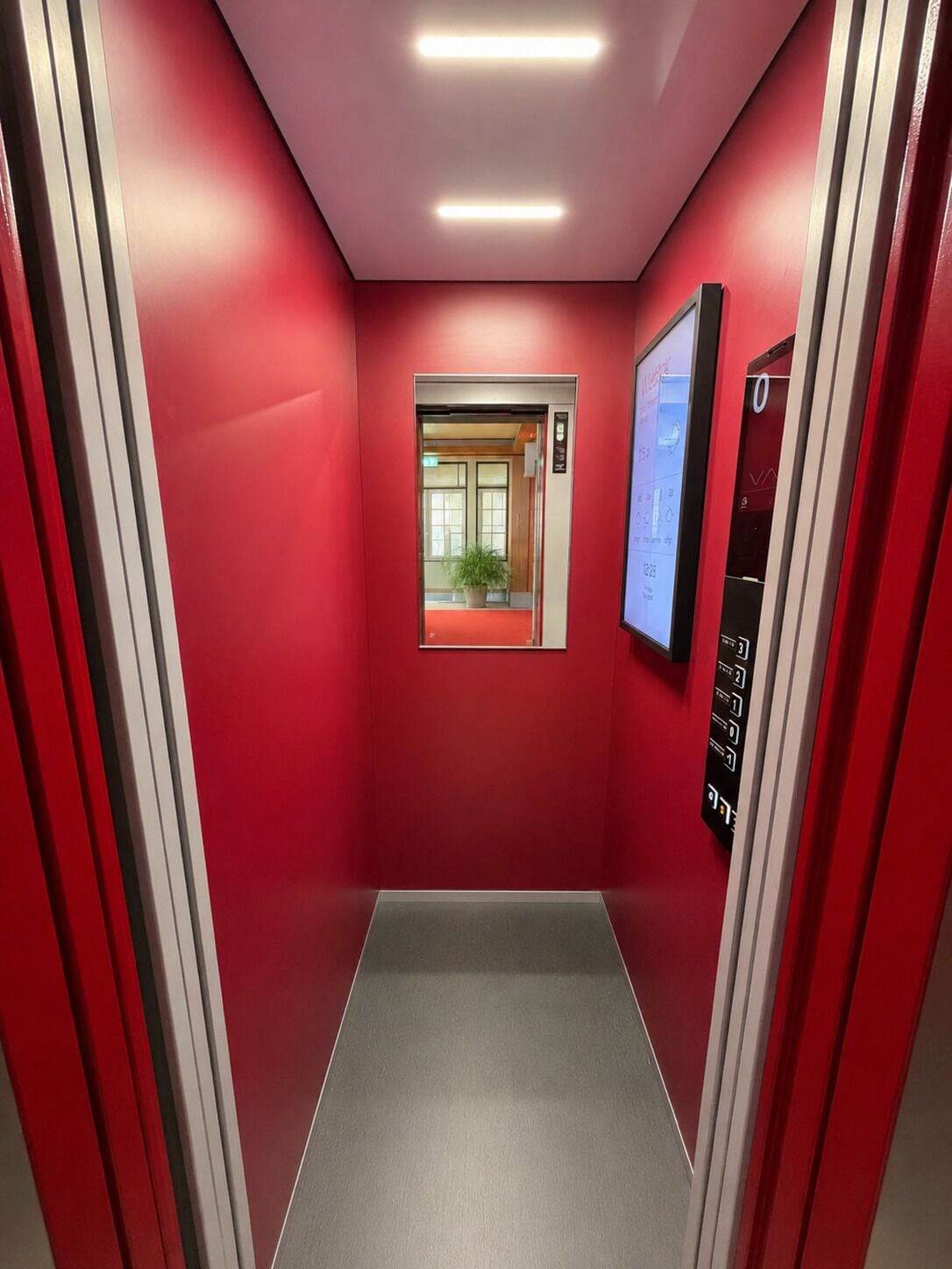 A red-walled elevator with a digital display, a potted plant, and a door with a sign.