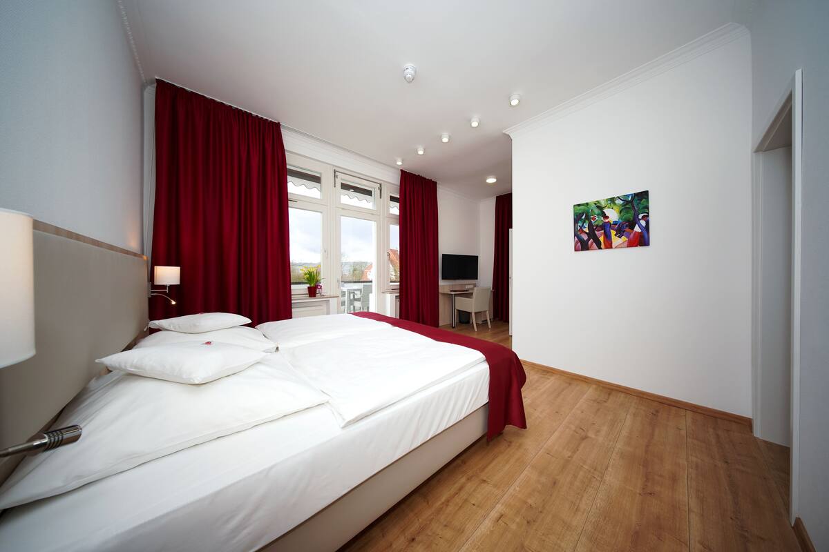 Quarto de hotel com uma cama grande, cortinas vermelhas, uma escrivaninha, uma cadeira e um quadro na parede.