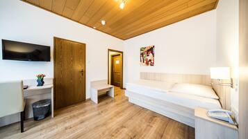 Una habitación de hotel moderna con techo de madera, televisor de pantalla plana montado en la pared, cama con cabecera blanca, mesita de noche con lámpara y un pequeño escritorio con un cesto de basura.