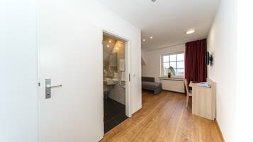 A modern, well-lit room with a wooden floor, a white door, a small kitchenette, a grey sofa, and a window with red curtains.