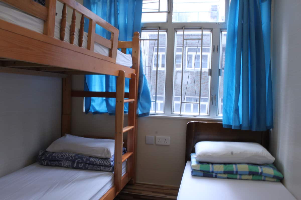 A dormitory room with bunk beds, a window with blue curtains, and a small table with a pillow.