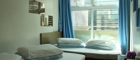 A hotel room with two beds, a window with blue curtains, and a view of buildings outside.