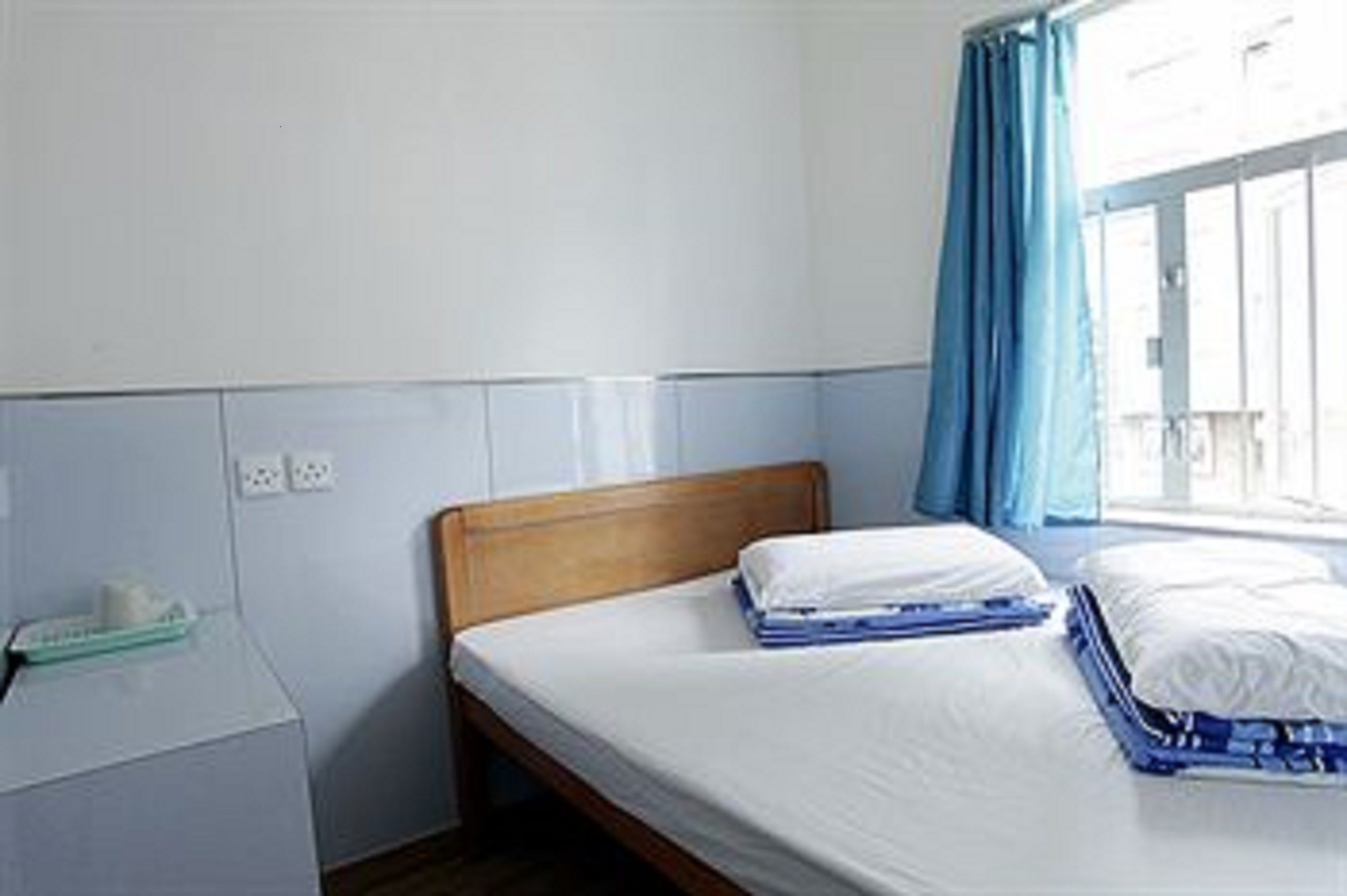 A single bed with a wooden headboard in a room with a white wall and a window with blue curtains.