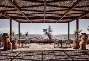 An outdoor seating area with tables and chairs, overlooking a body of water. - Numo Ierapetra Beach Resort Crete, Curio Collection Hilton (Ierapetra)