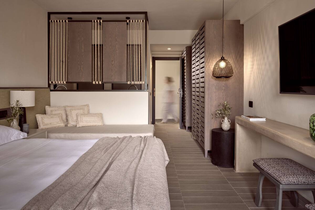 A modern hotel room with a large bed, a bedside table, a TV, and a seating area with a bench.