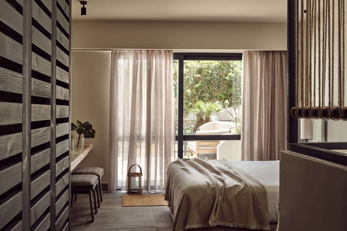 A bedroom with a bed, a bench, a small table, and a window with curtains.