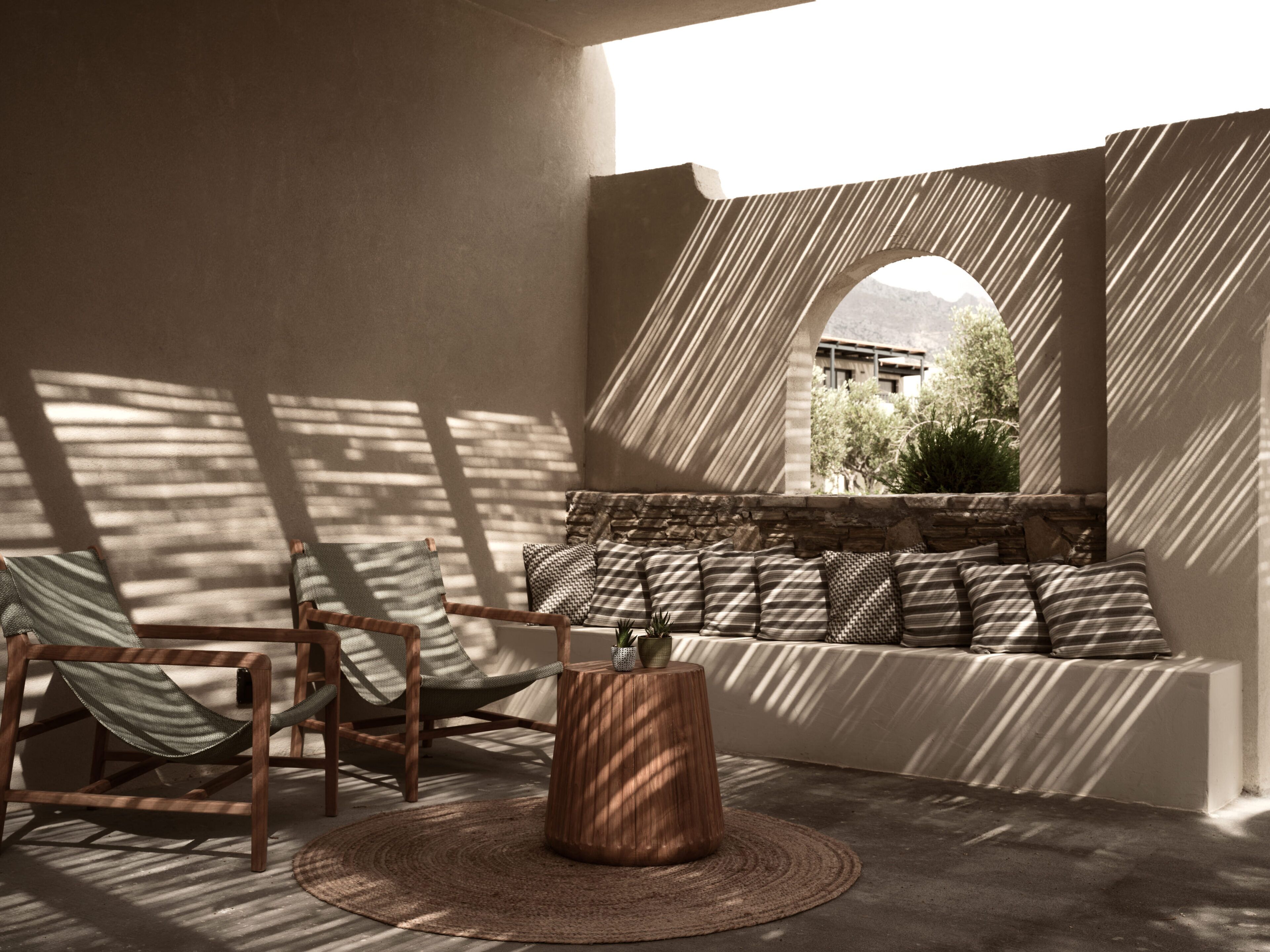 A sunlit seating area with a stone bench, two wooden chairs, and a woven rug.
