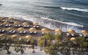 An aerial view of a beach with rows of sun loungers and thatched umbrellas, surrounded by trees. - Numo Ierapetra Beach Resort Crete, Curio Collection Hilton (Ierapetra)
