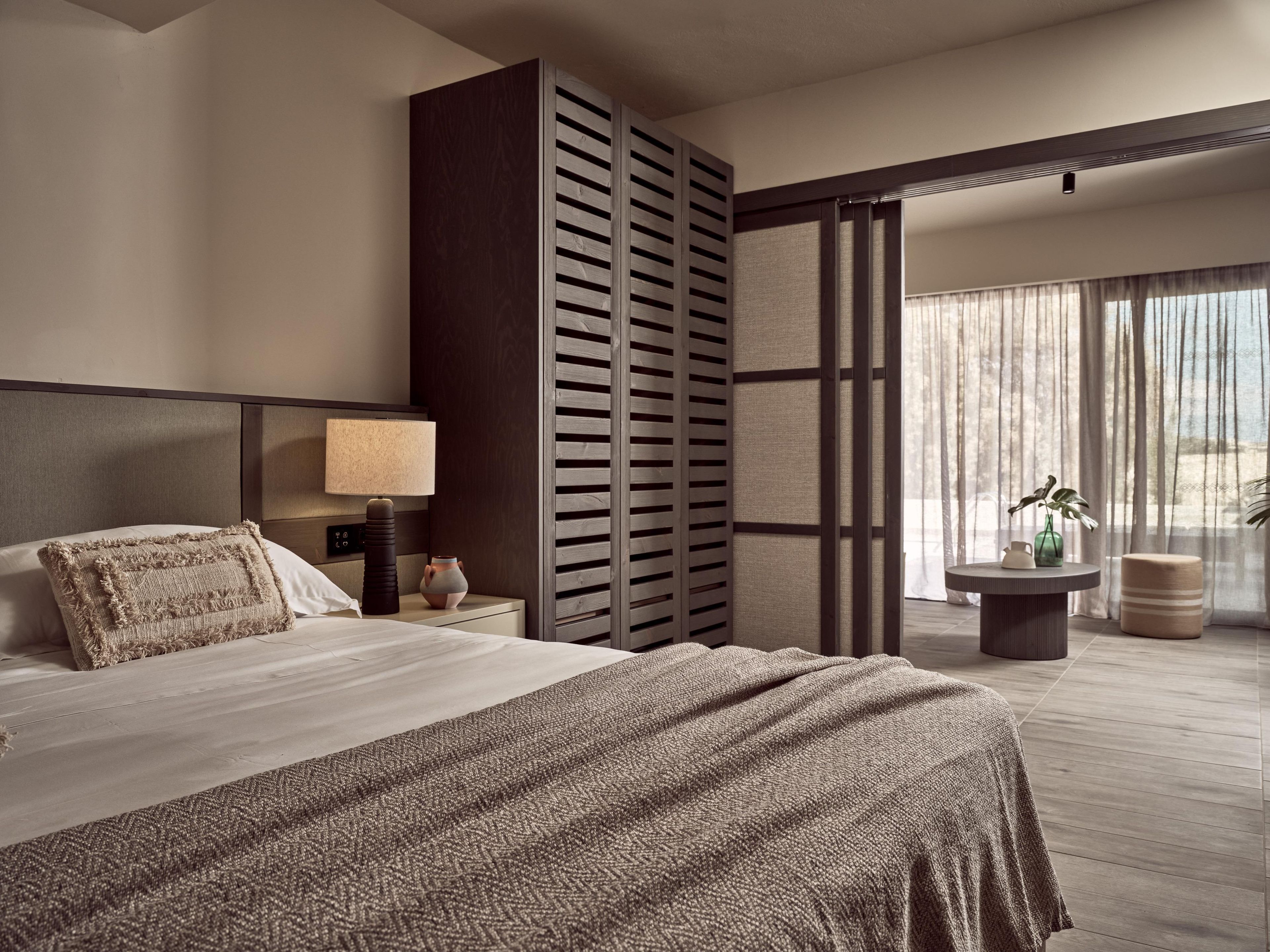 A modern hotel room with a large bed, a bedside table with a lamp, a decorative vase, and a view of the outdoors through sheer curtains.