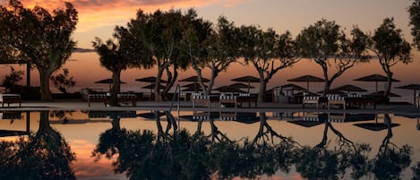 A serene waterfront setting with a reflective pool, lounge chairs, and umbrellas, reflecting the sunset sky.