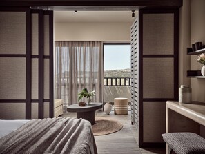 A hotel room with a bed, a round table with a plant, a view of the outdoors through a window, and a balcony with a railing. - Numo Ierapetra Beach Resort Crete, Curio Collection Hilton (Ierapetra)