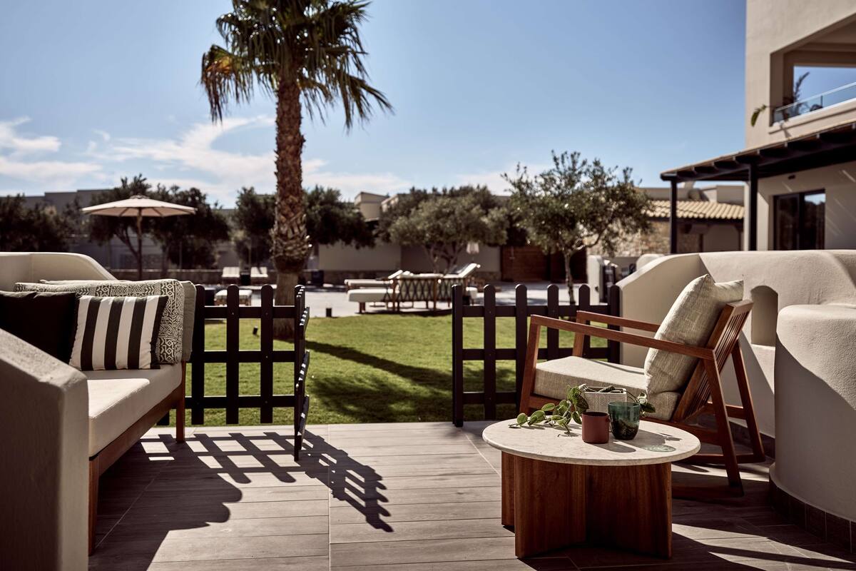 An outdoor patio area with a wooden deck, beige sofas, a round coffee table, and a palm tree.