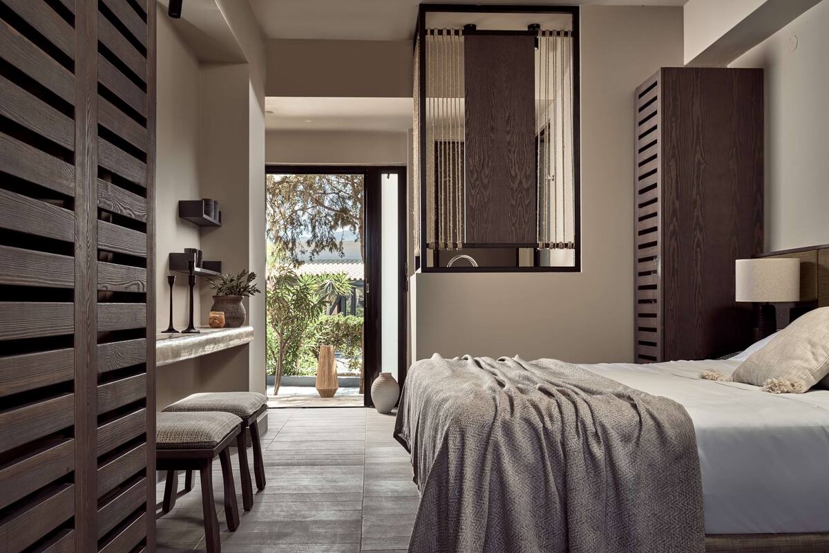 A modern bedroom with a large bed, a bedside table, a lamp, and a view of greenery outside.