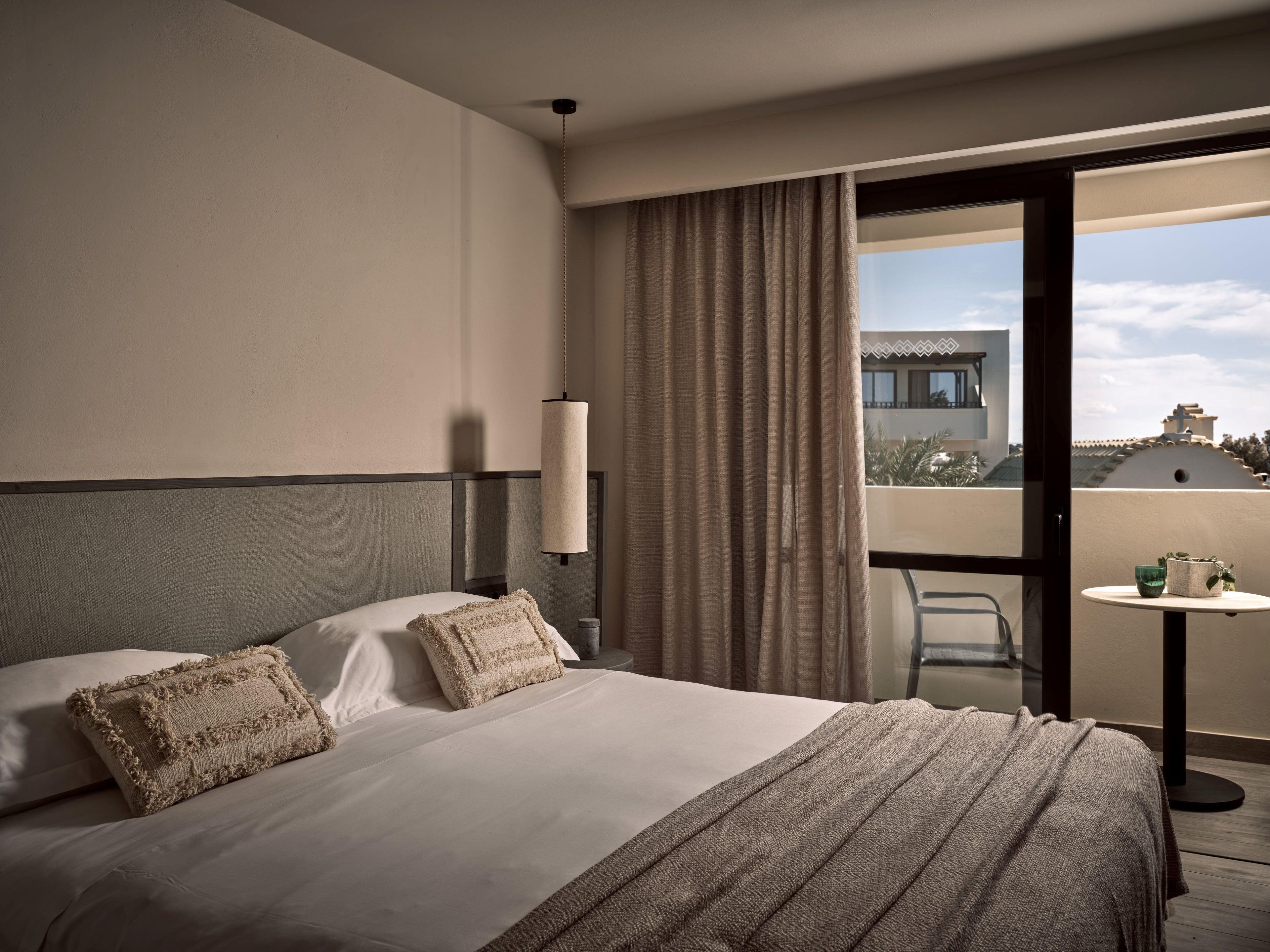 Une chambre d’hôtel avec un lit, une table de chevet, une lampe, une vue sur l’extérieur et un balcon avec une table et des chaises.