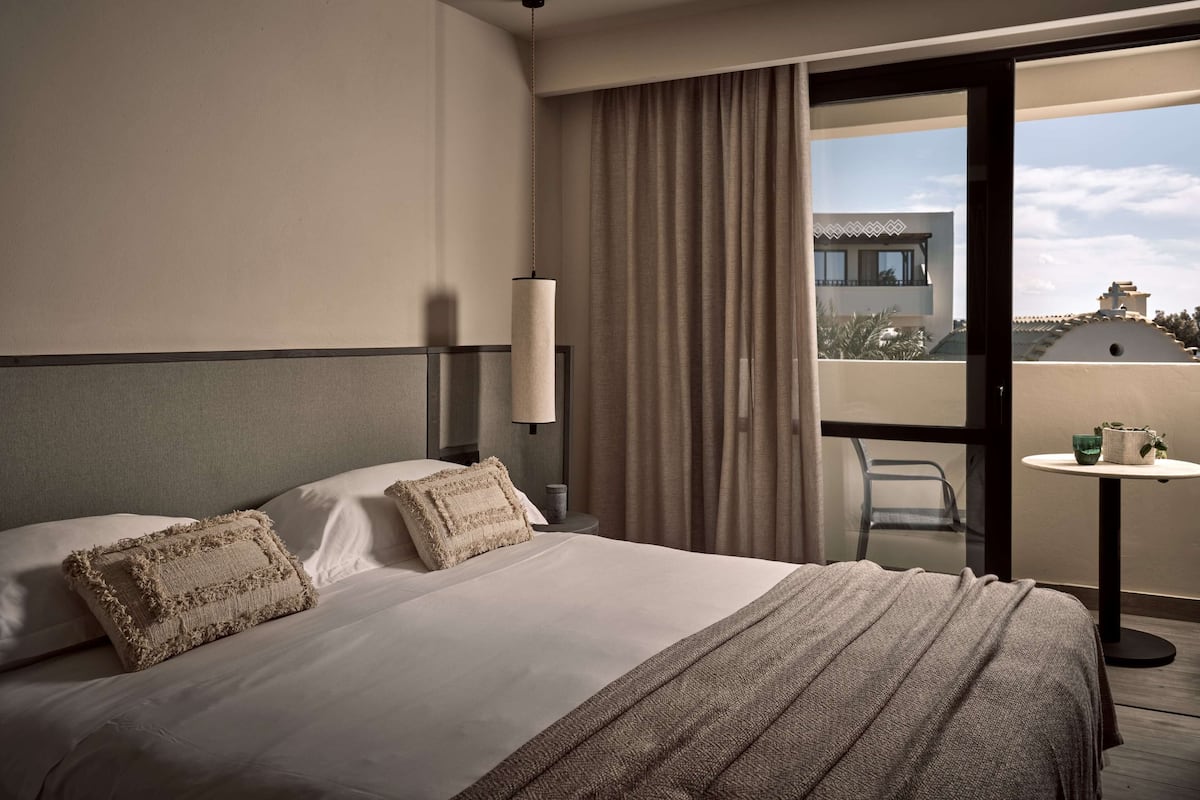 A hotel room with a bed, a bedside table, a lamp, a window with curtains, and a balcony with a table and chairs.