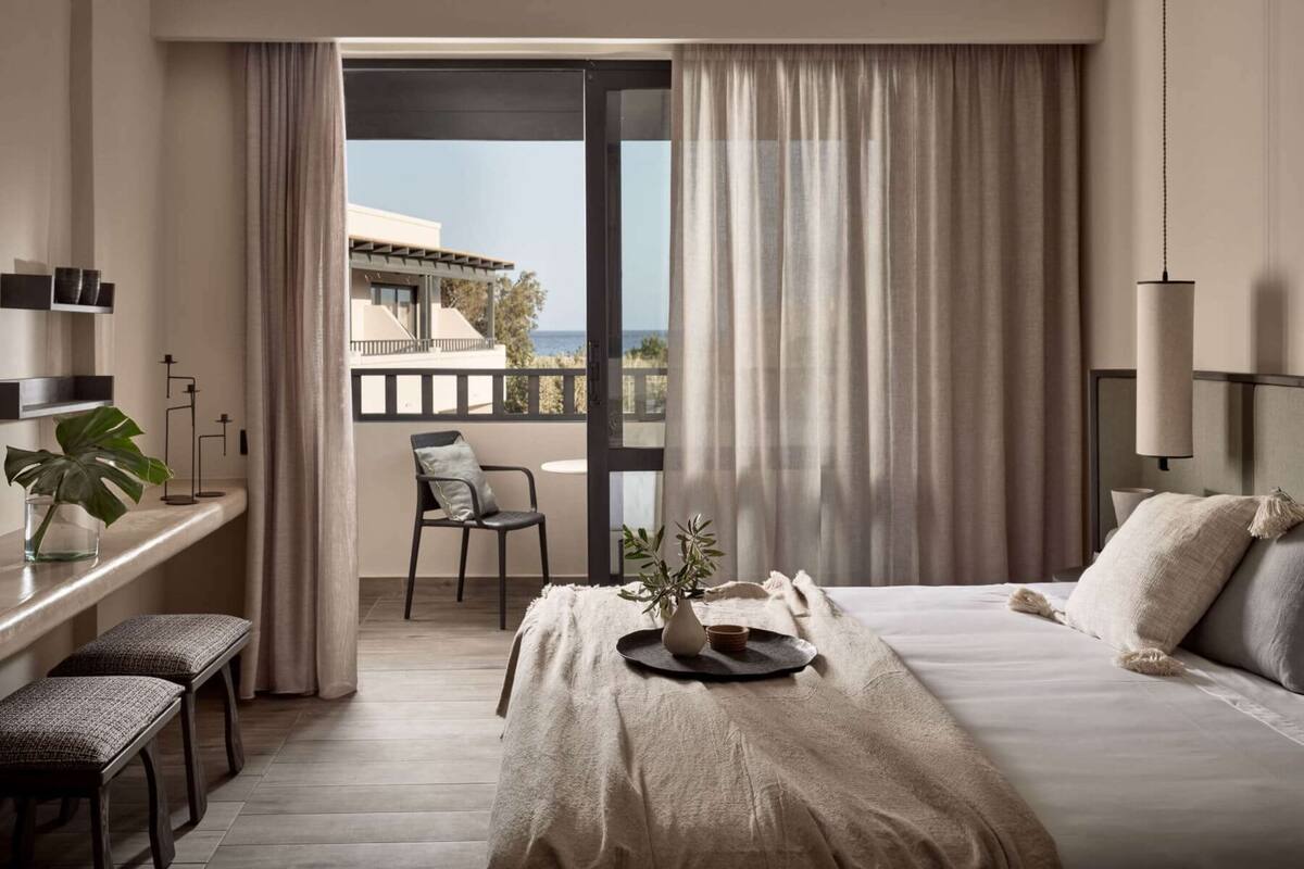 A hotel room with a bed, a desk with a chair, a potted plant, and a view of the sea through large windows.