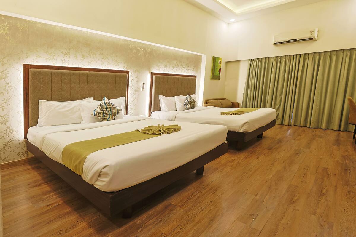 A hotel room with two beds, wooden flooring, a green curtain, and an air conditioning unit.
