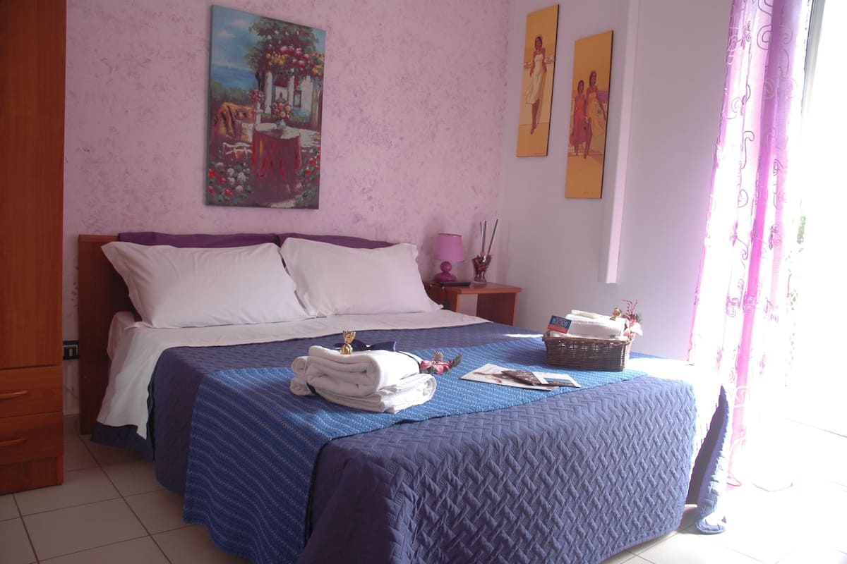 A neatly made bed with a blue quilt, a wooden nightstand with a lamp, and a painting on the wall.