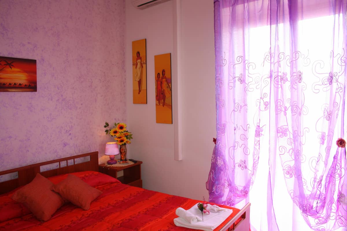 A bedroom with a red bedspread, a wooden headboard, a nightstand with a lamp and a vase of sunflowers, and a window with purple curtains.