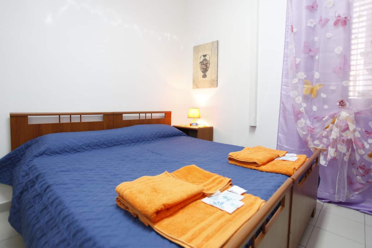 A bed with a blue bedspread, two folded orange towels, a wooden headboard, a nightstand with a lamp, and a wall-mounted picture.