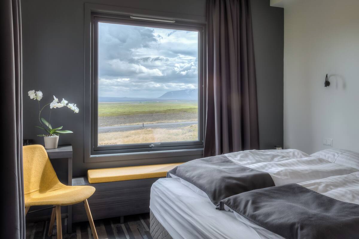Hotellihuone, jossa on suuri ikkuna, sänky, penkki, tuoli ja ruukussa oleva kasvi.