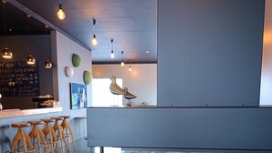 Et moderne barområde med en bardisk med glasplade, træbistrostole, hængende pendellamper og en synlig menutavle.