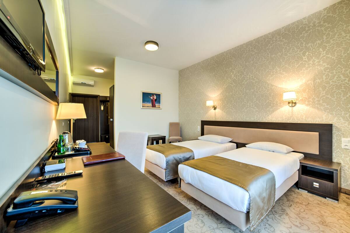 A hotel room with two beds, a desk with a lamp, a coffee maker, and a small table with a book and remote controls.
